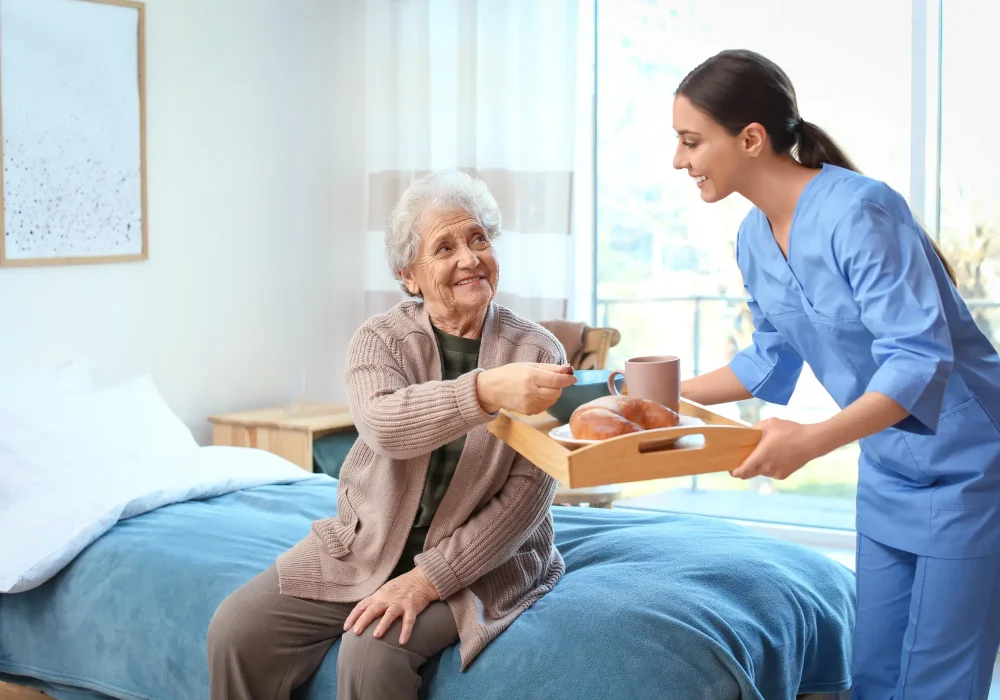 Elderly woman sitting on her bed while her nurse brings her food and drinks